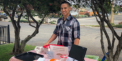 A person stands outdoors at a table with food samples and a sign, possibly related to nutrition or a dietetic intern project.