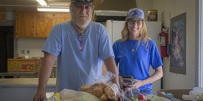 Dos personas de pie detrás de una mesa con alimentos, posiblemente en el entorno de una cocina comunitaria.