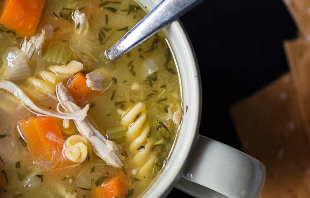 Un tazón de sopa de pollo con fideos, zanahorias, apio y hierbas, acompañado de galletas saladas.
