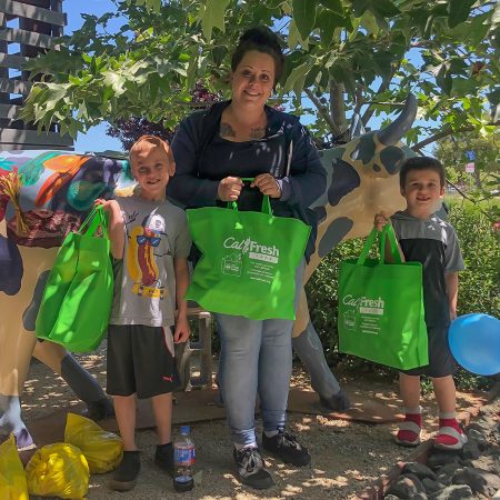 A woman and two children hold green CalFresh Food Benefits bags next to a painted cow sculpture outdoors.