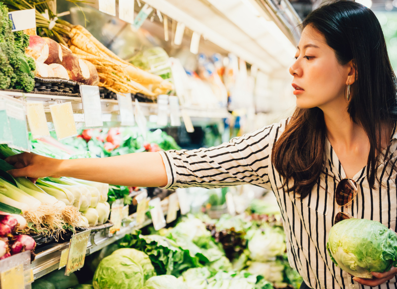 A woman grocery shopping, selecting fresh vegetables. Consider tips for saving money amid food inflation.