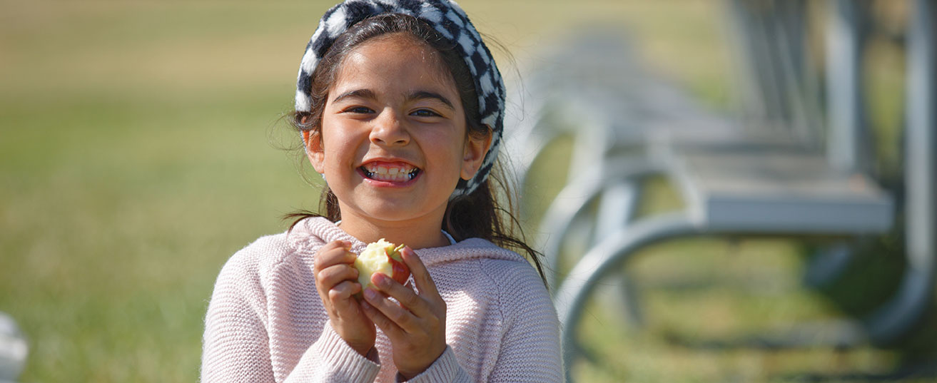 A smiling child is enjoying an apple outdoors, with benches in the background.