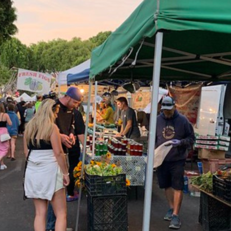 People shopping at an outdoor farmers' market with various stalls selling fresh produce, flowers, and jars of preserves. The market is bustling with activity, and there are trees and market lights.