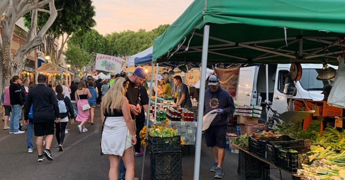 People shopping at an outdoor farmers' market with various stalls selling fresh produce, flowers, and jars of preserves. The market is bustling with activity, and there are trees and market lights.