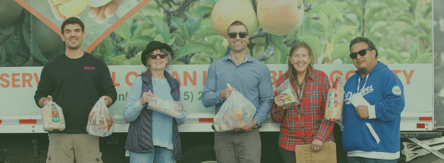 A group of people is standing in front of a truck holding bags with food items. SLO Food Bank is appropriate.