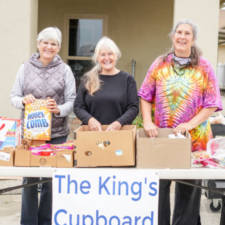 Three people are smiling behind a table with food items, possibly volunteering for local hunger relief organizations.