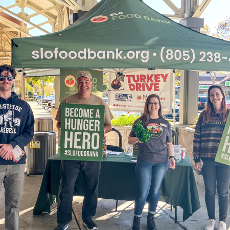 People are gathered under a SLO Food Bank tent for a turkey drive event, promoting hunger hero volunteering.