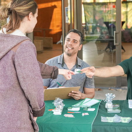 Two people seated at a table handing an item to a standing person.