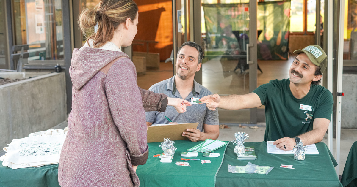 Two people seated at a table handing an item to a standing person.