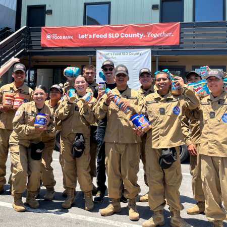A group of people in uniforms are smiling and holding packaged food items in front of a building with a food bank banner.