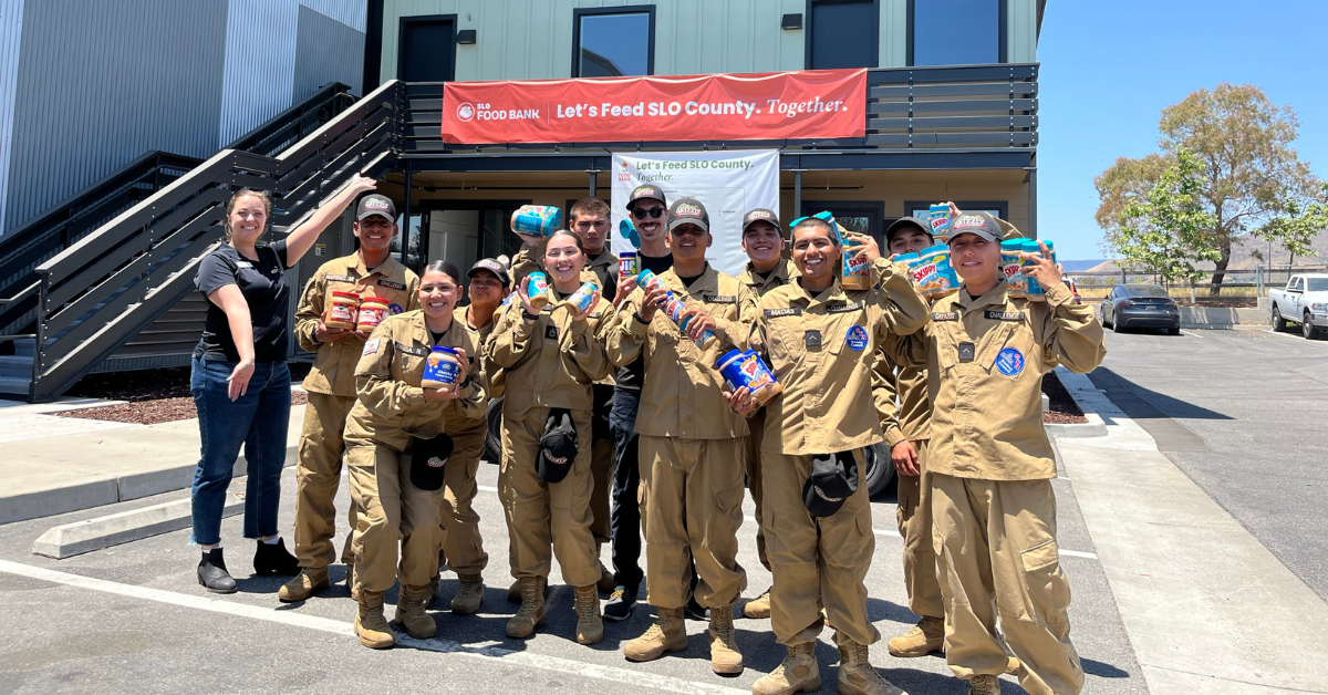 A group of people in uniforms are smiling and holding packaged food items in front of a building with a food bank banner.