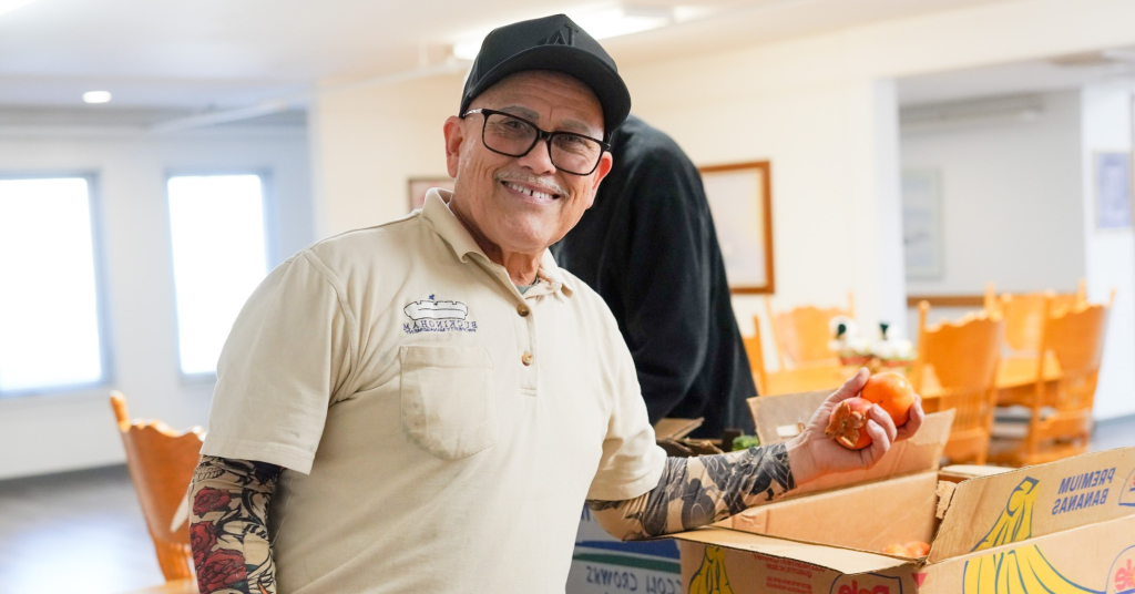 A person in a cap and glasses is smiling while holding tomatoes in a box. Senior food insecurity may be relevant.